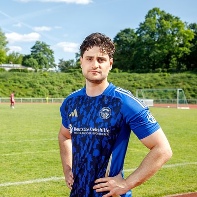 Junger Mann in blauem Fußballtrikot mit dem Logo der Deutschen Krebshilfe steht auf einem Fußballfeld mit grünen Bäumen im Hintergrund