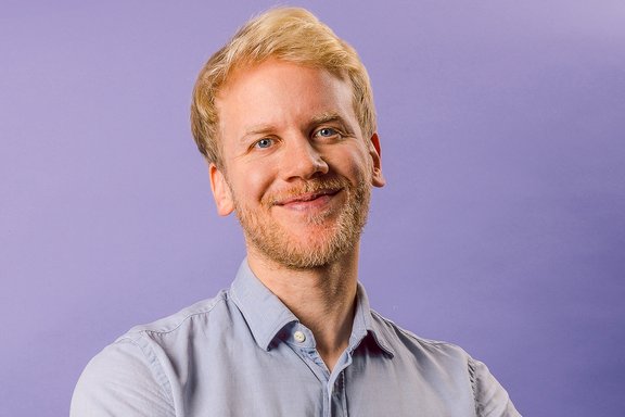 Mann mit hellblauem Hemd und hochgekrempelten Ärmeln steht vor lila Hintergrund mit verschränkten Armen und trägt eine Smartwatch am linken Handgelenk.