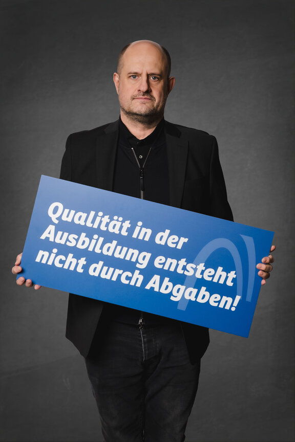 Eine Person steht vor einem neutralen grauen Hintergrund und hält ein großes blaues Schild mit der weißen Aufschrift: "Qualität in der Ausbildung entsteht nicht durch Abgaben"