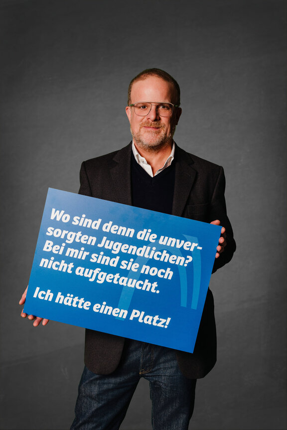 Eine Person steht vor einem neutralen grauen Hintergrund und hält ein großes blaues Schild mit der weißen Aufschrift: "Die Strafabgabe schwächt den Wirtschaftsstandort!"