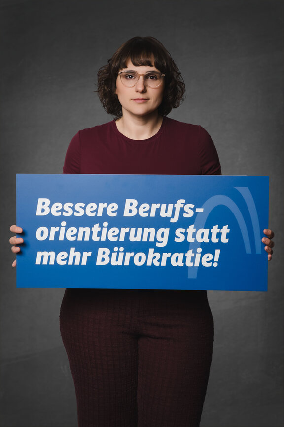 Eine Person steht vor einem neutralen grauen Hintergrund und hält ein großes blaues Schild mit der weißen Aufschrift: "Bessere Berufsorientierung statt mehr Bürokratie!"
