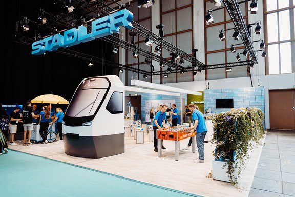 Messestand von Stadler mit großem Zugmodell, mehreren Personen in blauen Shirts und einem Tischfußballspiel