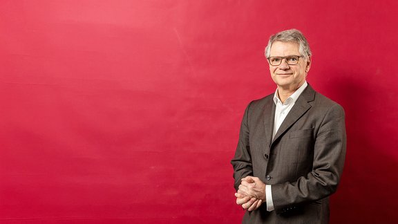 Mann in dunkelgrauem Anzug und weißem Hemd steht vor rotem Hintergrund, Hände vor dem Körper verschränkt