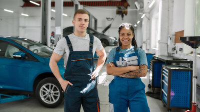 Zu sehen ist eine Autowerkstatt von innen. Im Zentrum des Bildes stehen zwei junge Personen, in T-Shirts und Latzhosen. Sie schauen freundlich und etwas stolz in die Kamera.