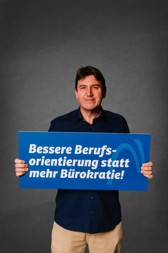 Eine Person steht vor einem neutralen grauen Hintergrund und hält ein großes blaues Schild mit der weißen Aufschrift: "Bessere Berufsorientierung statt mehr Bürokratie!"
