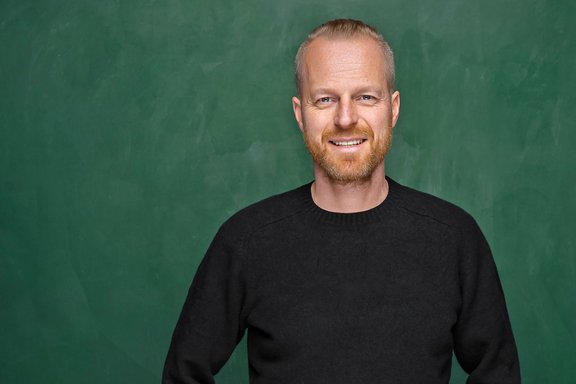 Mann mit kurzem Haar trägt schwarzen Pullover vor grüner Tafel
