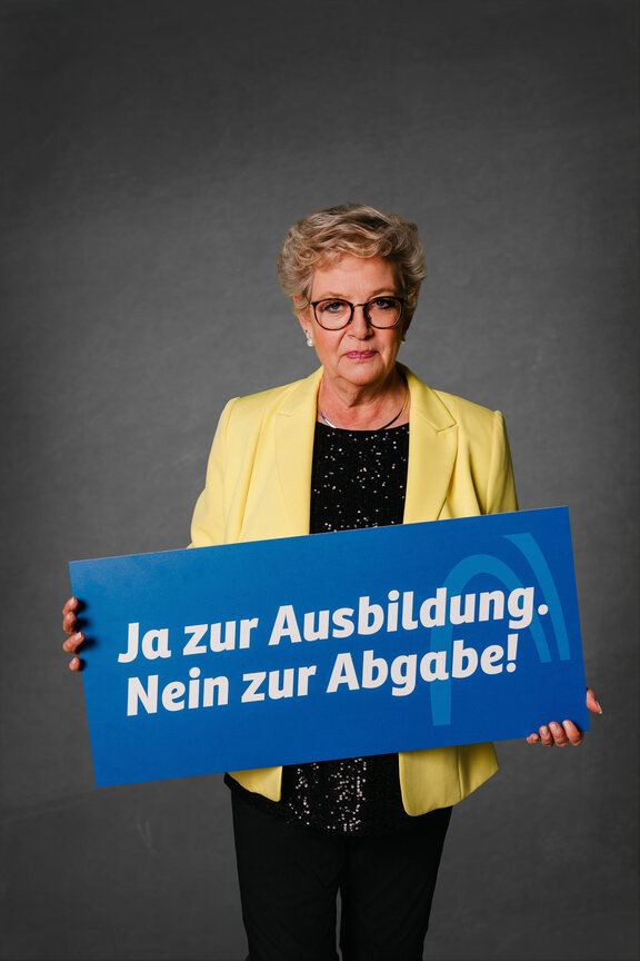 Eine Person steht vor einem neutralen grauen Hintergrund und hält ein großes blaues Schild mit der weißen Aufschrift: "Ja zu Ausbildung. Nein zur Abagabe!""