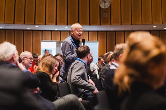 Zu sehen sind zahlreiche Menschen, die ein sitzendes Publikum Bilden. Eine Person von ihnen, ein älterer Herr mit weißen Haaren ist die einzige, die Steht. Der Mann hält ein Mikrofon und scheint eine Frage zu stellen.