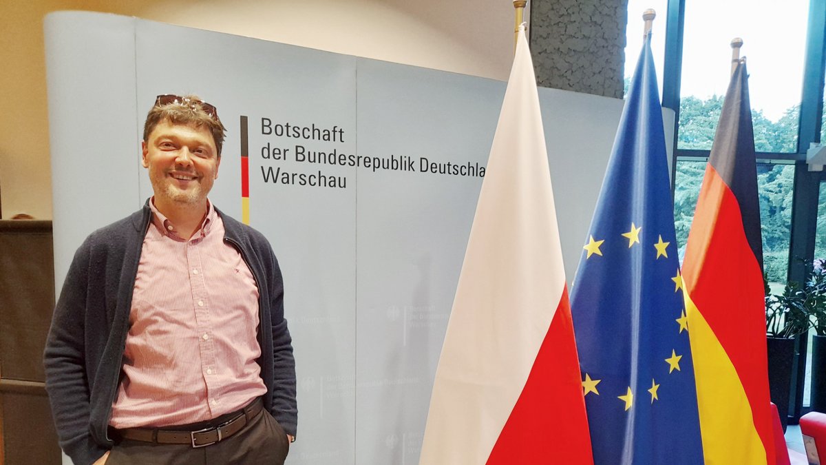 Mann mit rosa Hemd und dunklem Cardigan steht vor Wand mit Schrift 'Botschaft der Bundesrepublik Deutschland Warschau' neben den Flaggen von Polen, EU und Deutschland.