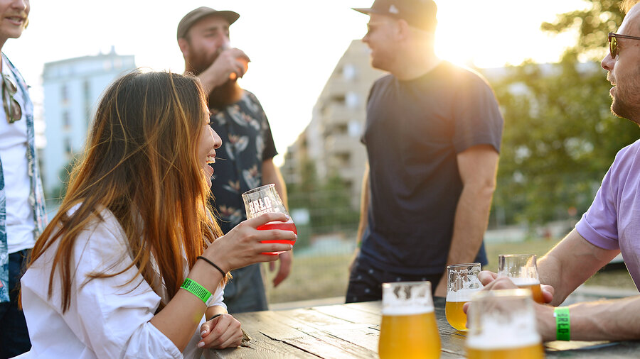 Menschen sitzen und stehen an einem Holztisch im Biergarten des BRLO Brwhouse mit Biergläsern bei Sonnenuntergang