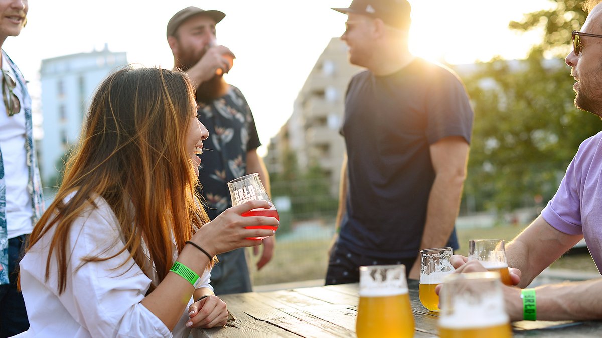 Menschen sitzen und stehen an einem Holztisch im Biergarten des BRLO Brwhouse mit Biergläsern bei Sonnenuntergang