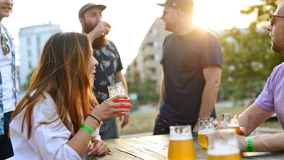 Menschen sitzen und stehen an einem Holztisch im Biergarten des BRLO Brwhouse mit Biergläsern bei Sonnenuntergang