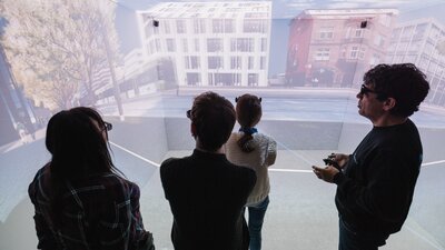 Vier Personen stehen in einem Raum, auf dessen Wände die Umgebung auf einer Straße projeziert wird.