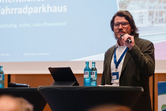 Zu sehen ist ein Mann mir Bart und langen Haaren in einem dunkelgrünen Sakko. Er steht auf einer Bühne und spricht in ein Mikrofon zu Publikum.