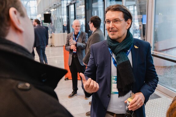 Man Blickt von hinten über die Schulter eines Mannes, der sich mit einem Mann im Sakko unterhält.