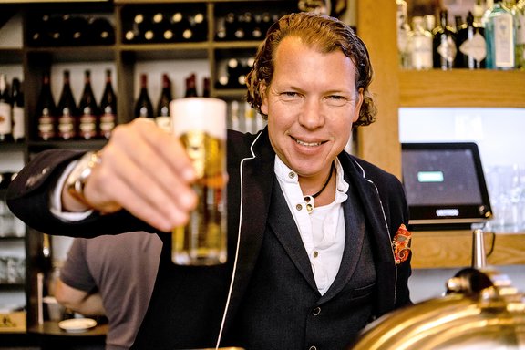 Ein Mann in einem schwarzen Anzug mit weißem Hemd und einer roten Tasche hält ein Glas Bier in der Hand. Im Hintergrund sind Weinflaschen und eine Bar zu sehen.