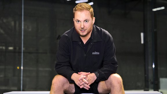 Mann sitzt mit verschränkten Händen auf einer Bank, trägt schwarze Jacke mit Aufschrift 'mitte' und Shorts.