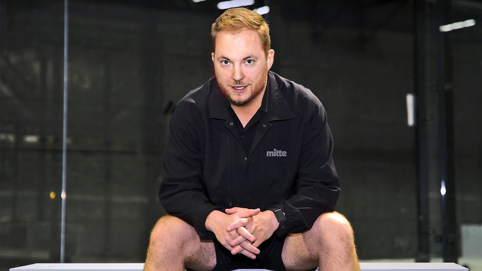 Mann sitzt mit verschränkten Händen auf einer Bank, trägt schwarze Jacke mit Aufschrift 'mitte' und Shorts.