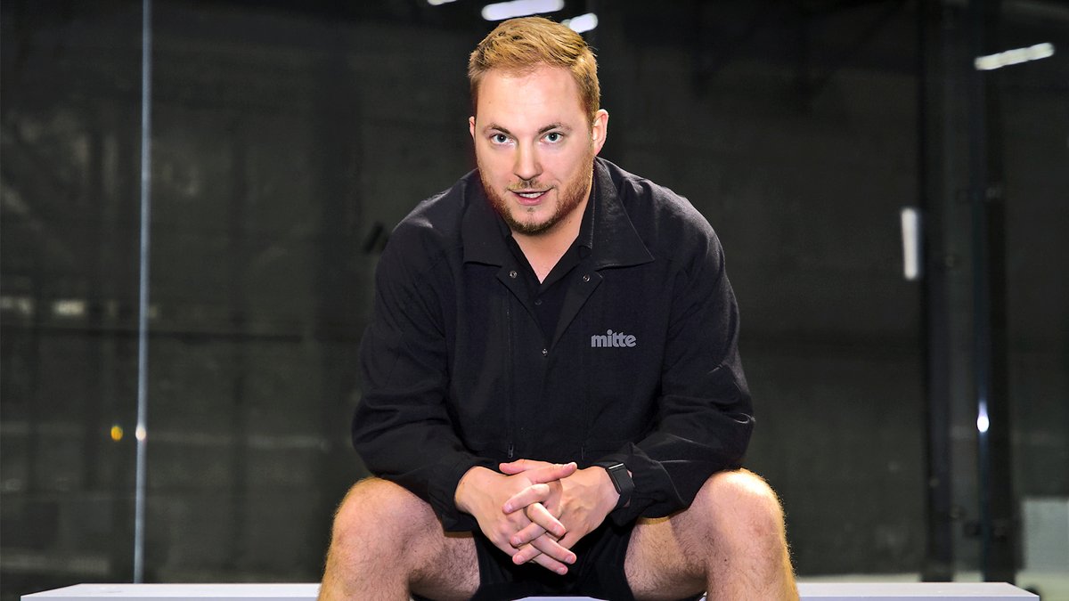 Mann sitzt mit verschränkten Händen auf einer Bank, trägt schwarze Jacke mit Aufschrift 'mitte' und Shorts.