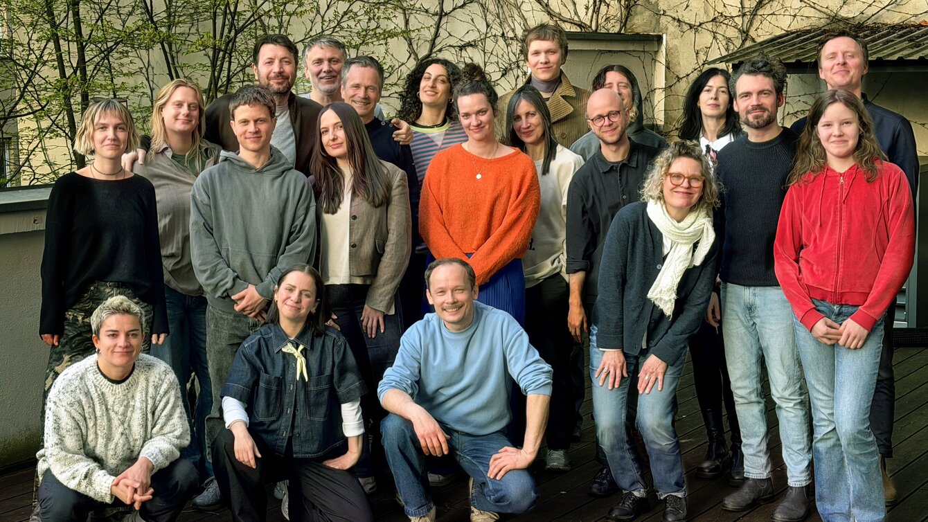 Gruppenaufnahme mehrerer Personen aus einem Unternehmen, die für ein Teamfoto nebeneinander vor einem schlichten Hintergrund posieren; das Team gehört zu einer Agentur für Audio Branding und Motion Design.