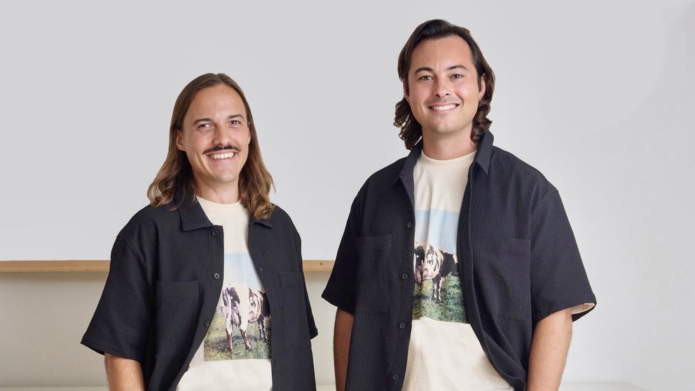 Zwei Personen stehen nebeneinander, beide tragen beige Hosen, weiße T-Shirts mit Tiermotiv und schwarze Kurzarmhemden, vor weißem Sideboard und heller Wand.