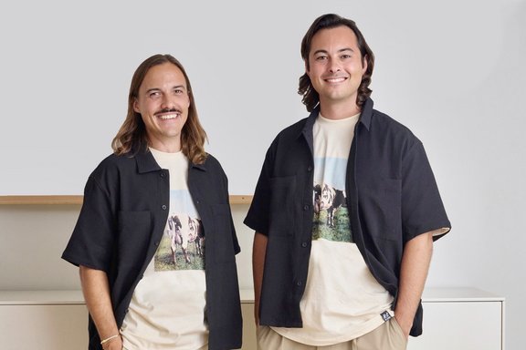Zwei Personen stehen nebeneinander, beide tragen beige Hosen, weiße T-Shirts mit Tiermotiv und schwarze Kurzarmhemden, vor weißem Sideboard und heller Wand.