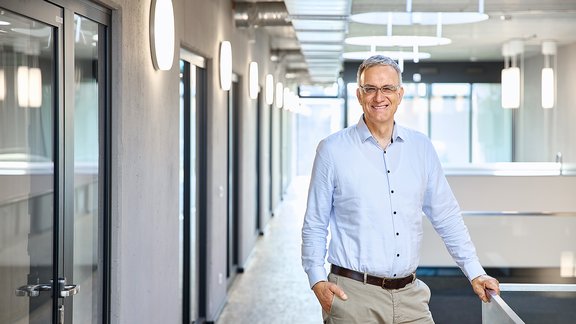 Mann mit grauen Haaren, trägt ein hellblaues Hemd und beige Hose, steht entspannt in einem modernen Büroflur mit großen Fenstern und runden Deckenleuchten.