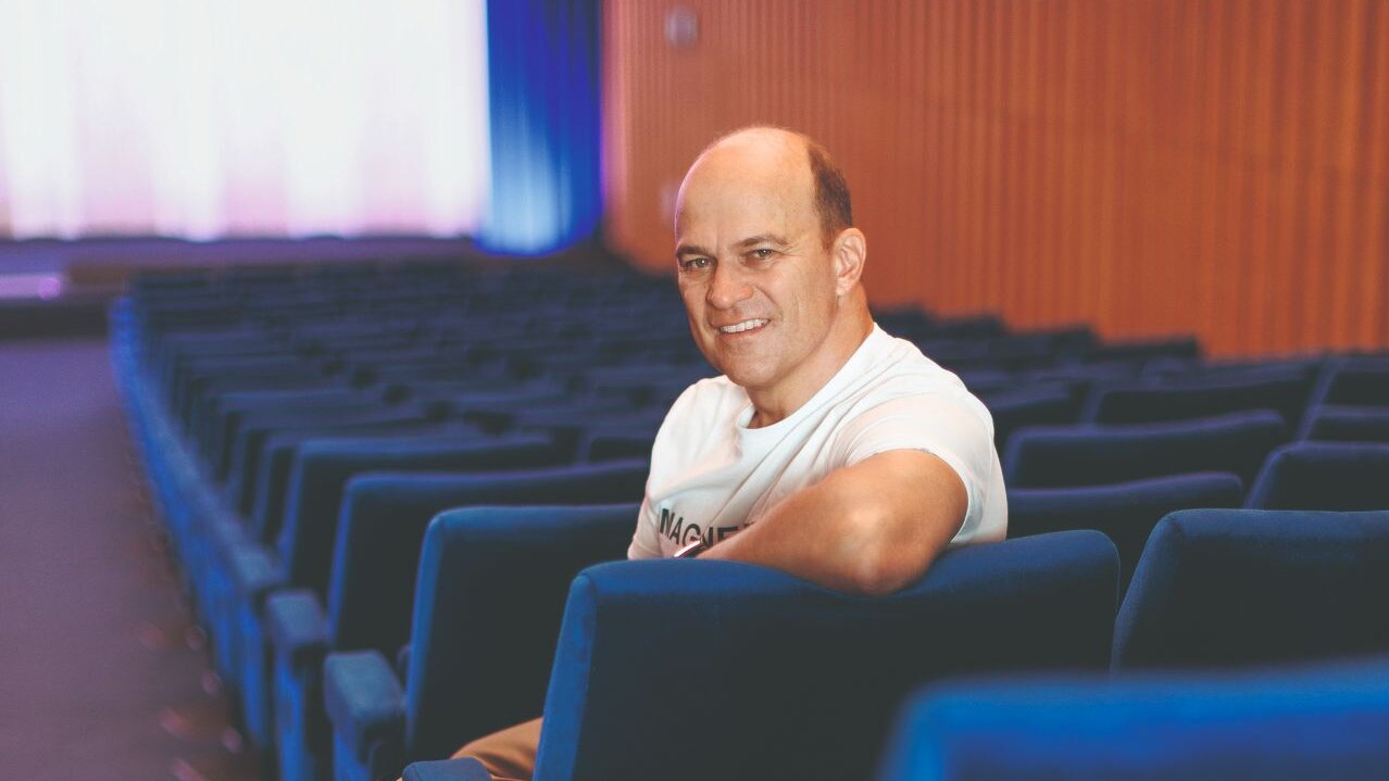 Zu sehen ist ein Mann mit weißem Shirt, der auf einem Sitz in einem leeren Kino sitzt und freundlich in die Kamera blickt.