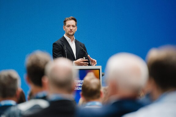Zu sehen ist ein junger Mann hinter einem Redepult, davor sieht man die Köpfe der Publikums.