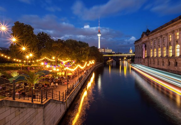 Abendliche Uferpromenade in Berlin mit beleuchteten Bäumen und Sitzplätzen links, Fluss in der Bildmitte, Fernsehturm und beleuchtete Gebäude im Hintergrund rechts.