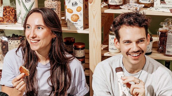 Eine junge Frau und ein junger Mann lachen beide fröhlich und essen KoRo Snacks vor einem Regal mit KoRo-Produkten.