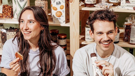 Zwei Personen sitzen vor einem Regal mit verpackten Lebensmitteln, eine hält ein Stück Brot, die andere eine verpackte Riegelstange.