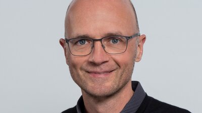Auf dem Bild ist ein Mann mit Glatze und Brille zu sehen, der in die Kamera lächelt.