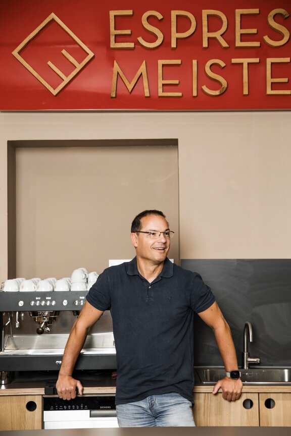 Auf dem Bild sieht man einen Mann, der an einer Theke lehnt. Über ihm hängt ein Schild mit der Aufschrift "Espressomeisterei".