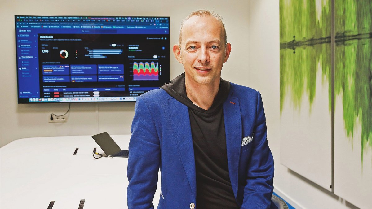 Mann lehnt an einem Tisch in einem Meetingraum mit einem Fernseher im Hintergrund.