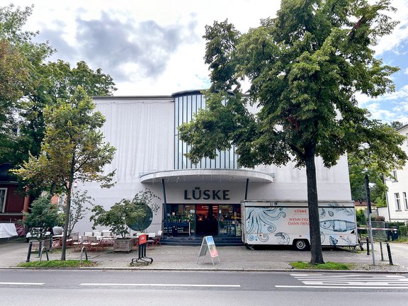 Gebäude mit dem Schriftzug 'LÜSKE' über dem Eingang, davor Bäume, ein Lieferwagen mit Muster und Text 'LÜSKE WENN FISCH DANN FRISCH' und ein Bürgersteig mit Sitzgelegenheiten