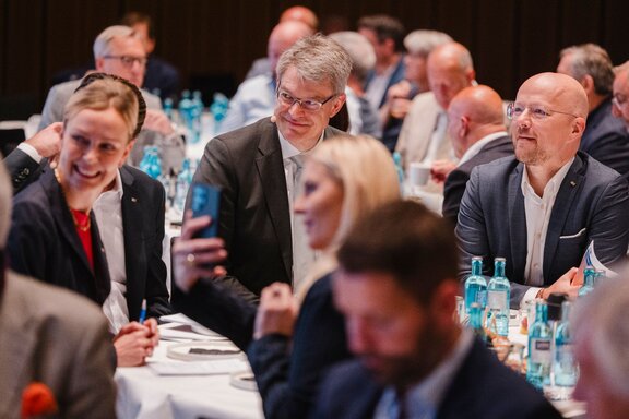 Zu sehen sind viele Personen, die an Tischen sitzen und auf eine Bühne zu schauen scheinen.
