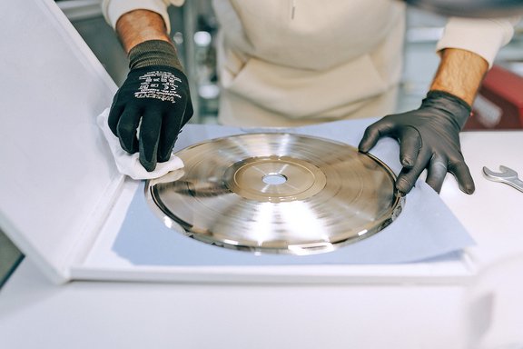 Person mit schwarzen Handschuhen reinigt eine glänzende Schallplatte auf einem weißen Gerät