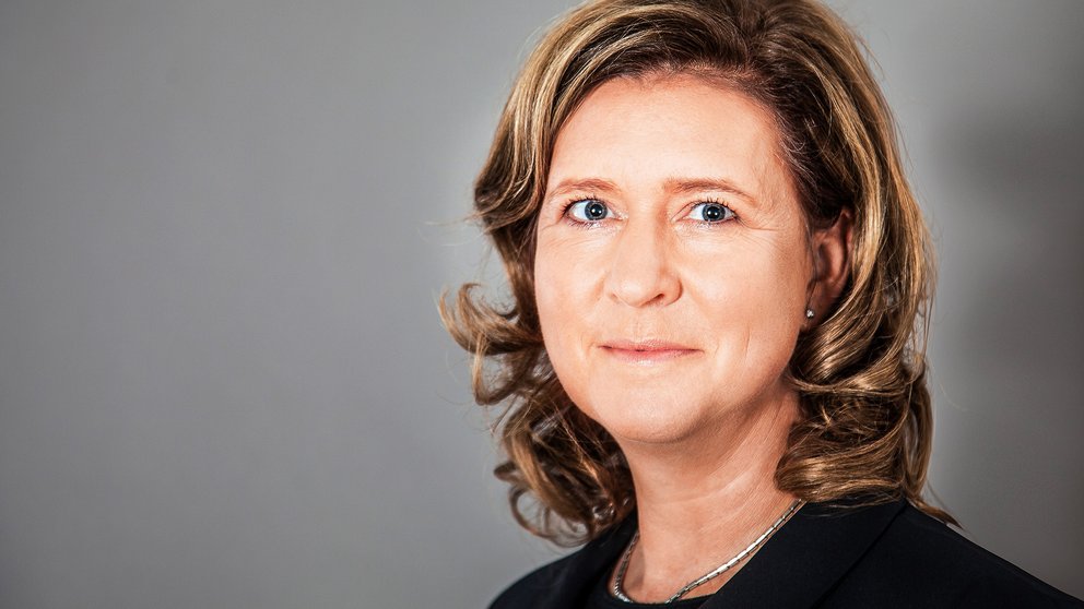 Frau mit schulterlangen, gewellten blonden Haaren, trägt schwarzen Blazer und silberne Halskette vor grauem Hintergrund.
