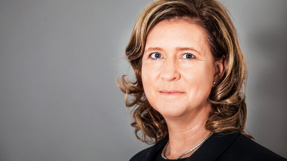 Frau mit schulterlangen, gewellten blonden Haaren, trägt schwarzen Blazer und silberne Halskette vor grauem Hintergrund.
