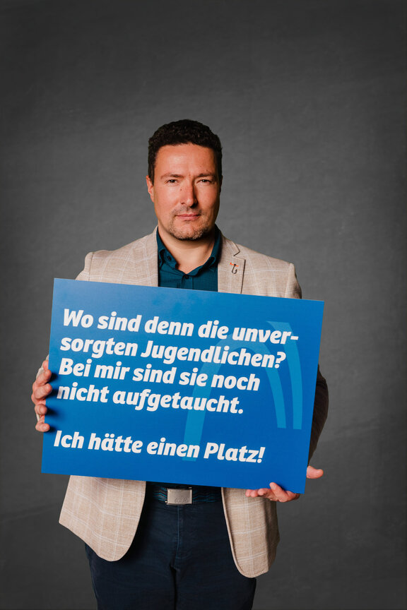 Eine Person steht vor einem neutralen grauen Hintergrund und hält ein großes blaues Schild mit der weißen Aufschrift: "Wo sind denn die unversorgten Jugendlichen? Bei mir sind sie noch nicht aufgetaucht. Ich hätte einen Platz!"