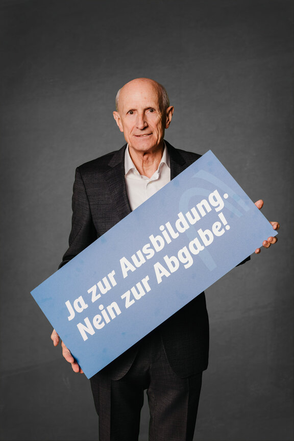 Eine Person steht vor einem neutralen grauen Hintergrund und hält ein großes blaues Schild mit der weißen Aufschrift: "Ja zur Ausbildung. Nein zur Abgabe!"