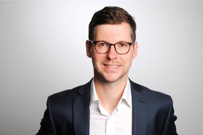Portrait Patrick Bergmann, mit kurzen braunen Haaren, Brille und dunklem Anzug lächelt vor hellem Hintergrund