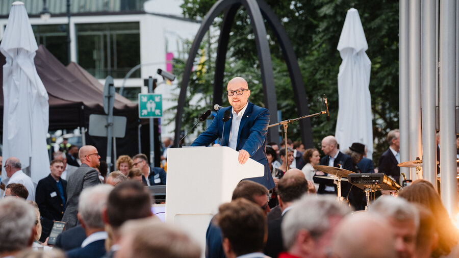 Zu sehen ist ein Redner, welcher an einem weißen Redepult steht. Die Rede findet unter freiem Himmel statt, während zahlreiche Personen im Publikum stehen.