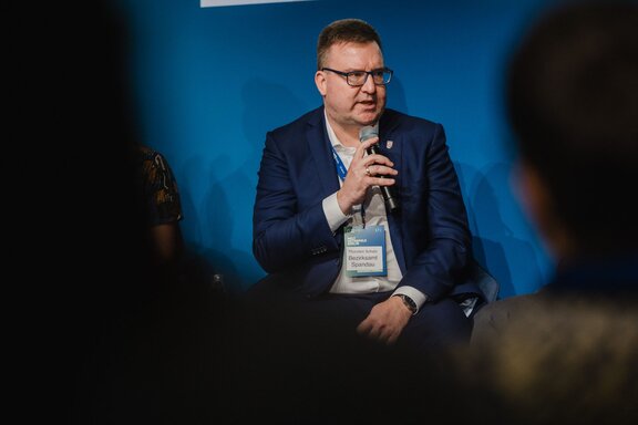 Ein Mann spricht sitzend auf einer Bühne vor Publikum.