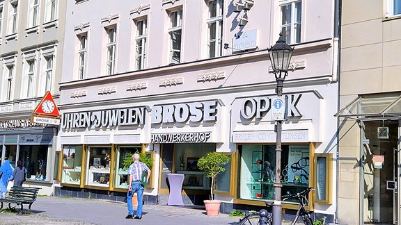 Fassade eines mehrstöckigen Gebäudes mit Geschäften für Uhren, Schmuck und Optik, davor Pflasterstraße und Fahrradständer.