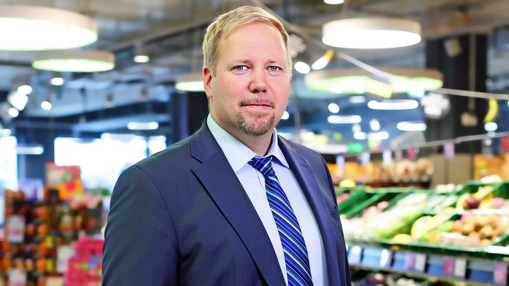Ein Mann im Anzug steht in einem Supermarkt-Gang mit Lebensmittelregalen. 