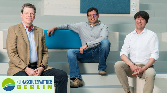 Drei Männer sitzen auf einer hellblauen Treppe und schauen freundlich in die Kamera. 