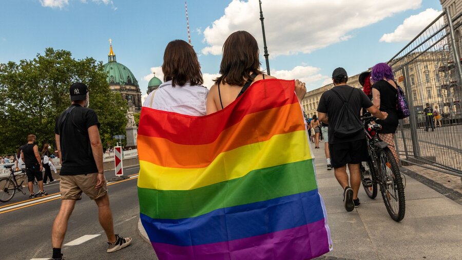 Zu sehen sind zwei Personen, die eine Regenbogenfahne um sich gehüllt haben und eine Straße entlang laufen.