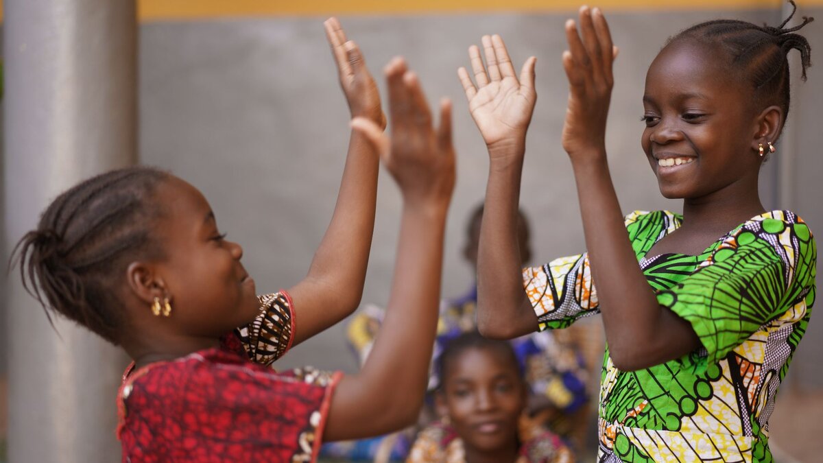 Zu sehen sind zwei Mädchen in bunten Kleidern, welche lachen und gegenseitig in die Hände klatschen.